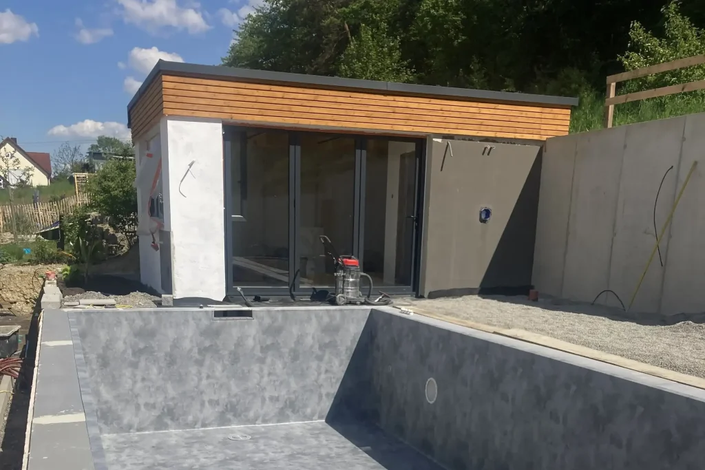 Unfertige Poolanlage mit modernem Gartenhaus im Bau, sichtbare Leitungen und grauer Betonpool ohne Wasser.