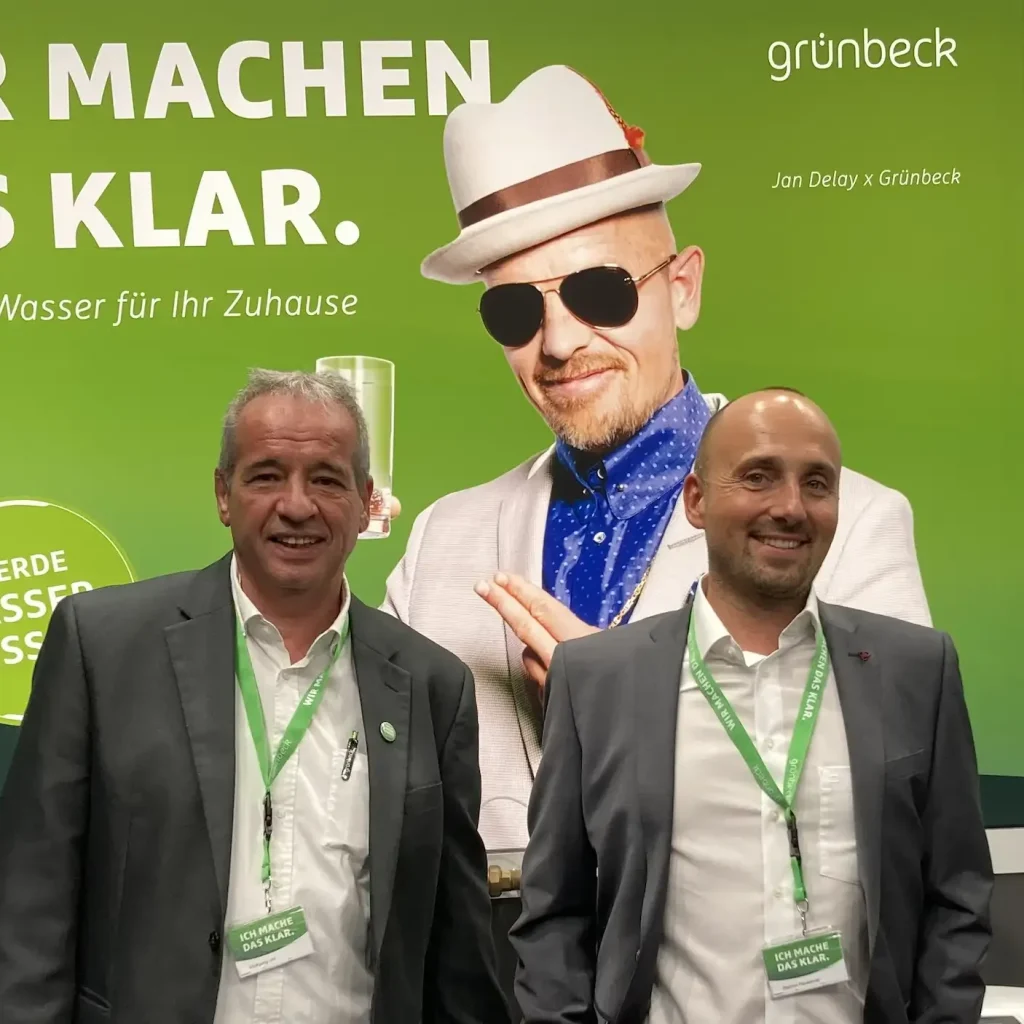 Grünbeck-Mitarbeiter am Messestand vor grünem Hintergrund mit Werbeslogan und Foto von Werbebotschafter Jan Delay.