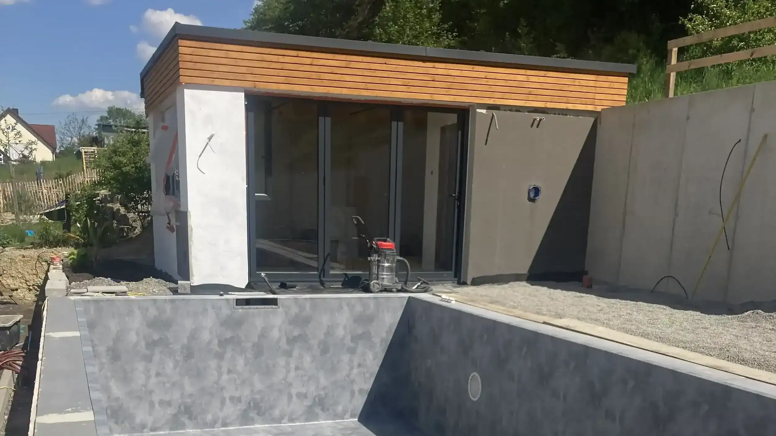 Modernes Poolhaus mit Holzlamellen und Schiebetüren; davor unfertiger Pool in der Sanierungsphase.