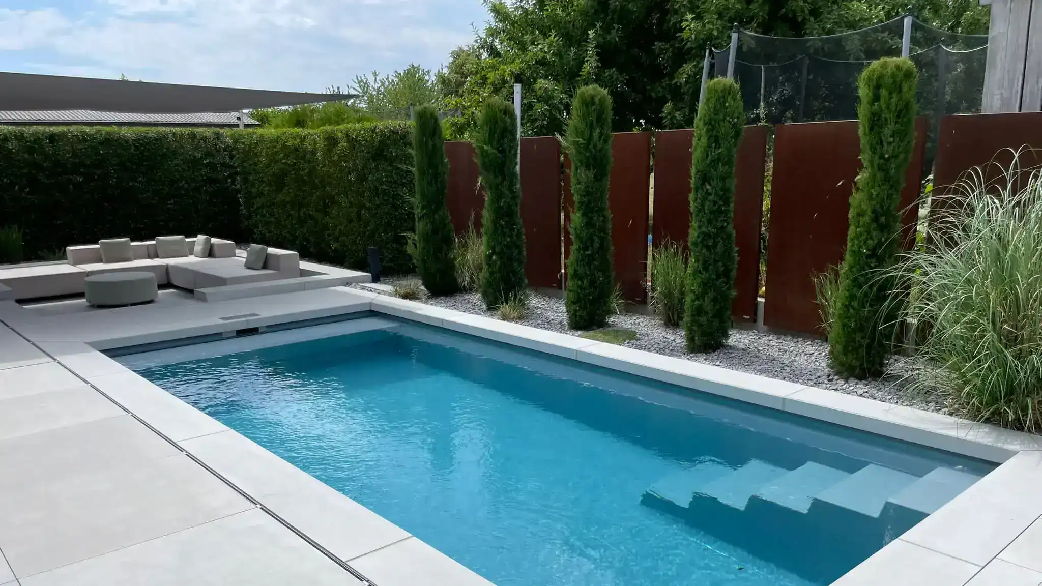 Moderner rechteckiger Gartenpool mit Treppe; helle Terassenplatten; im Hintergrund Sitzlounge, Hecke und Sichtschutz.