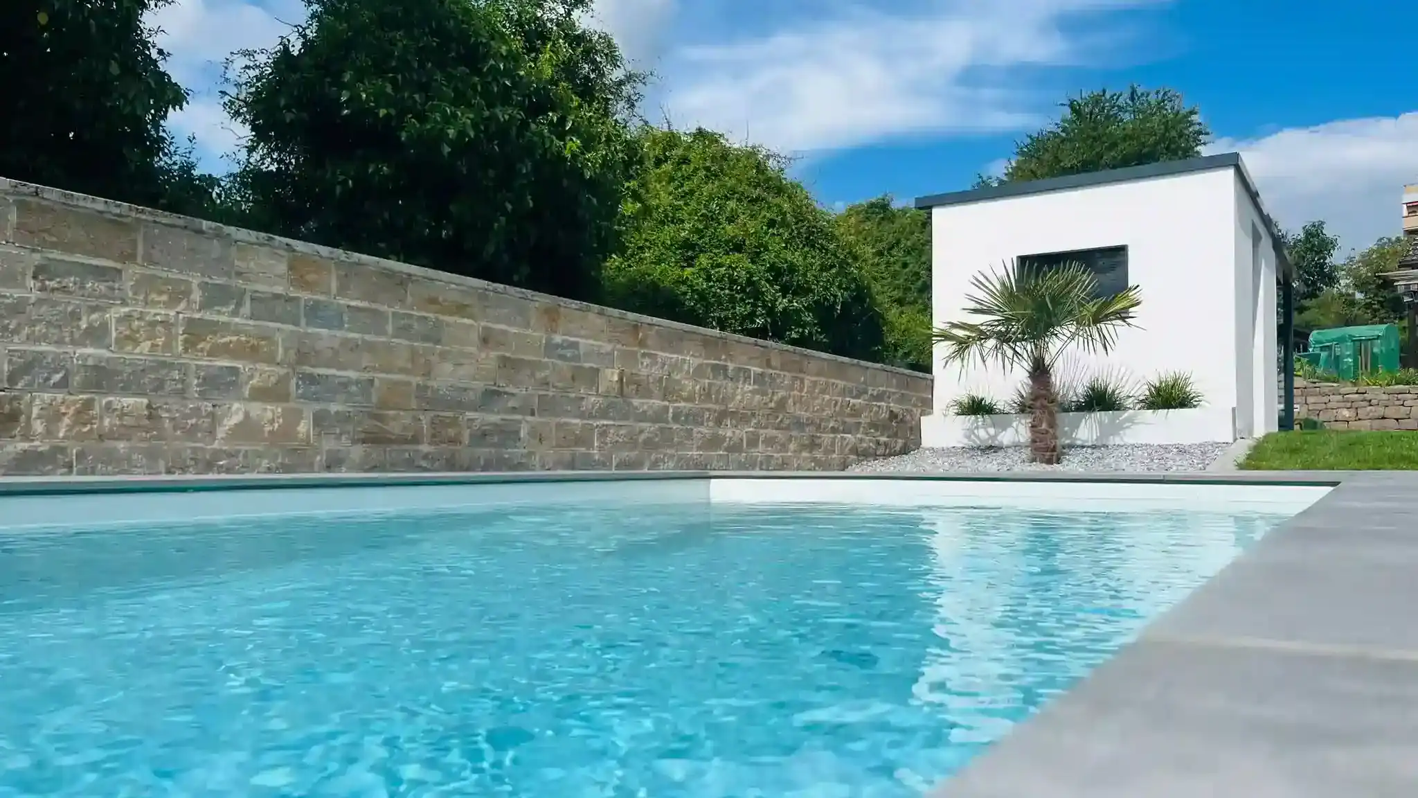 Outdoorpool mit blauem Wasser; Naturstein-Stützmauer entlang des Pools; im Hintergrund weißes Poolhaus mit Palme.