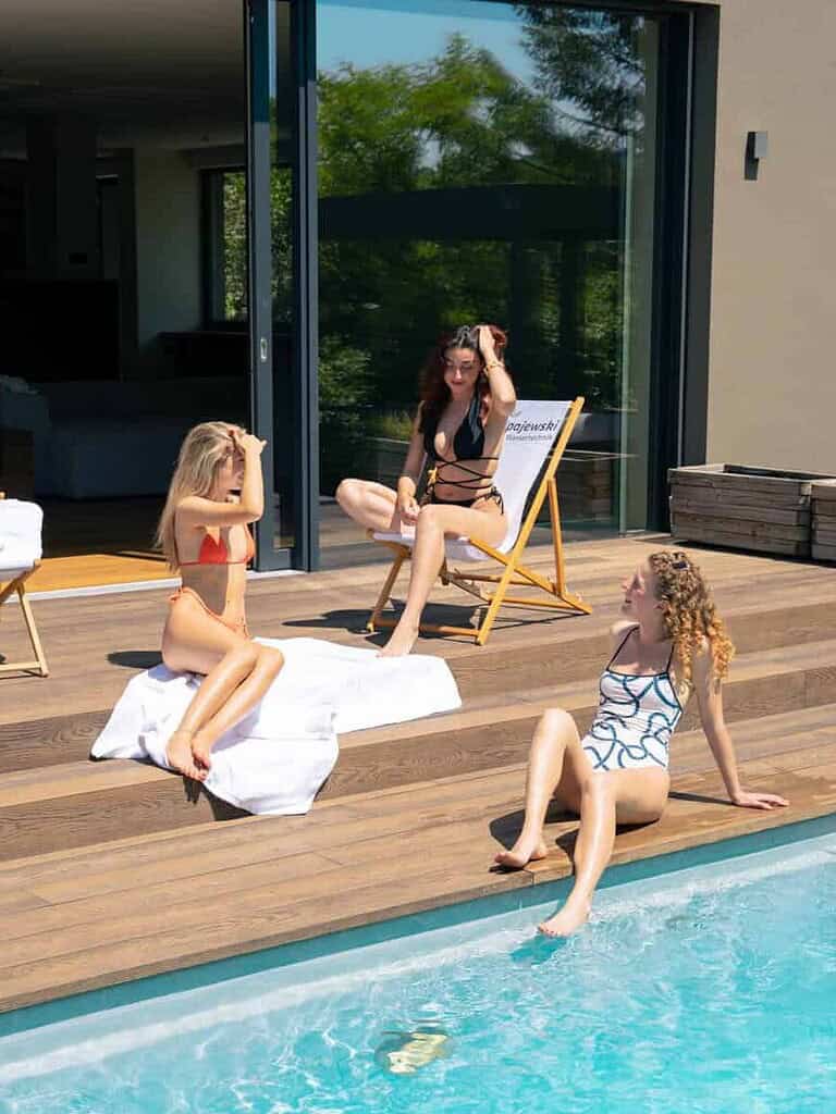 Drei Frauen entspannen auf einer Holzterrasse am privaten Pool; eine Frau hat ihren linken Fuß im Wasser.