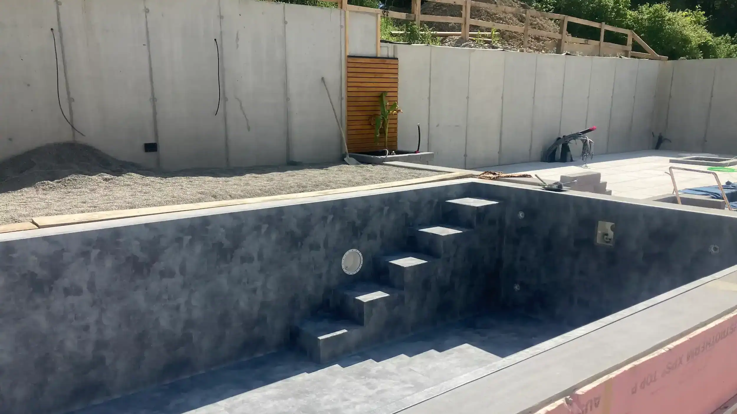 Unbefüllter grauer Betonpool mit integrierter Treppe, im Garten im Bauzustand mit sichtbarer Betonmauer.